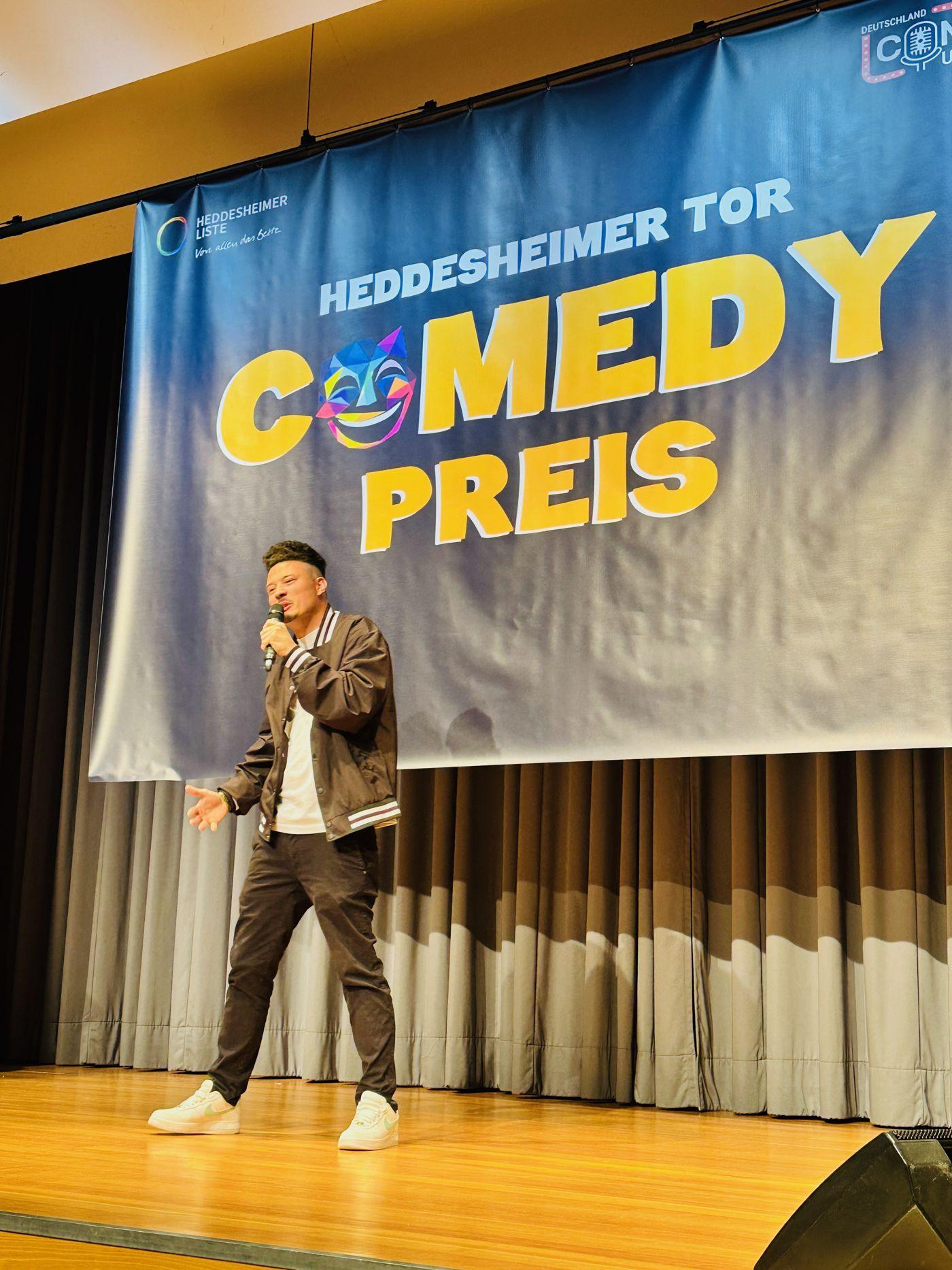 Comedian in Jacke auf der Bühne, dahinter das Banner „Heddesheimer Tor Comedypreis“