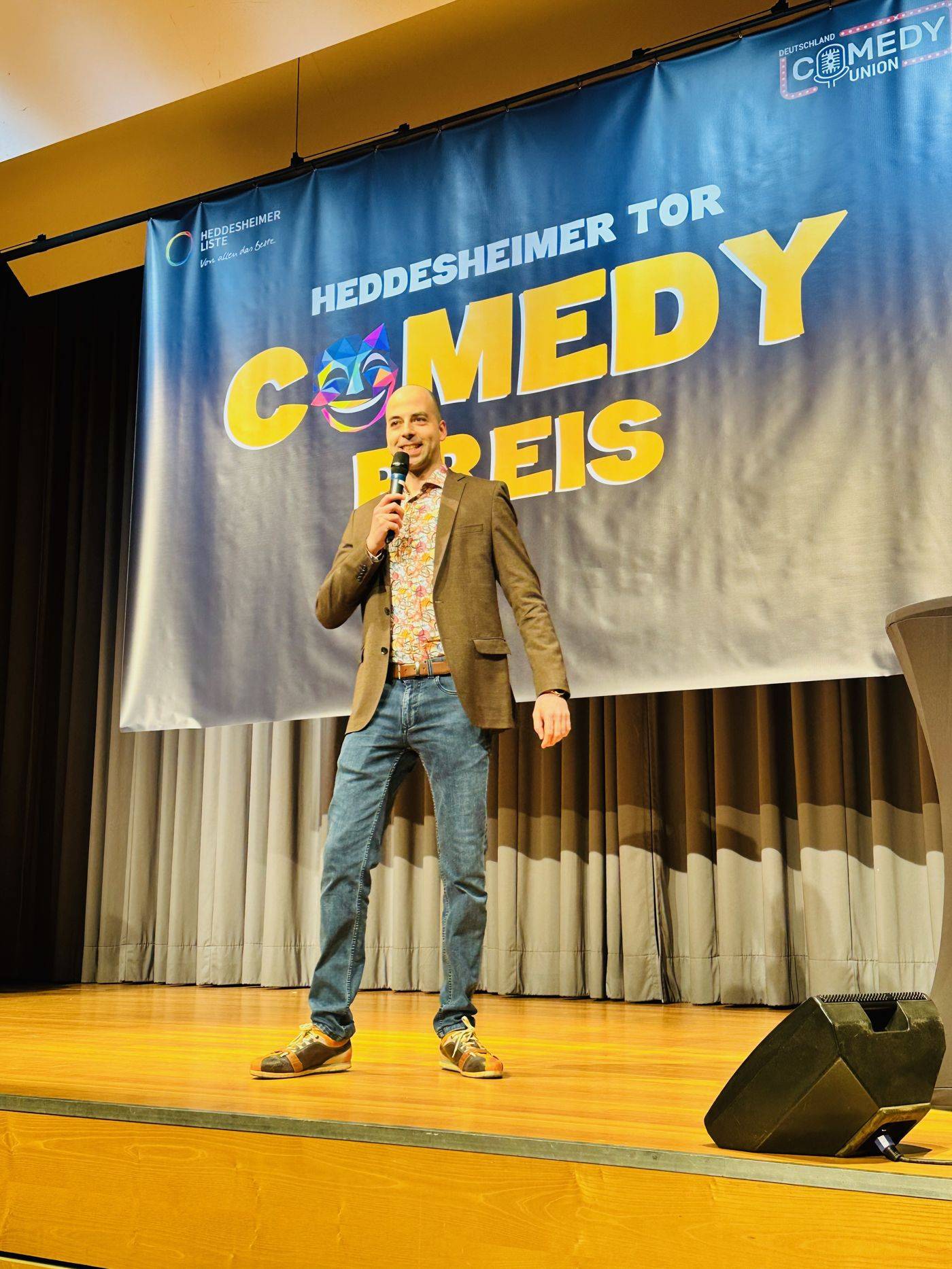 Comedian im Sakko auf der Bühne vor dem Banner